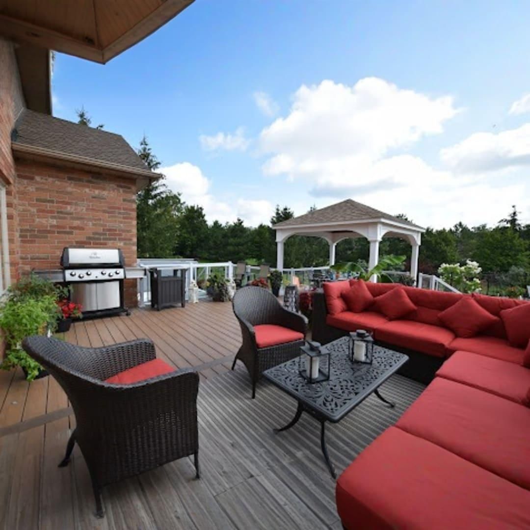 A wooden deck features a red sectional sofa, a black wicker armchair, and a coffee table, with a grill and a gazebo nearby.