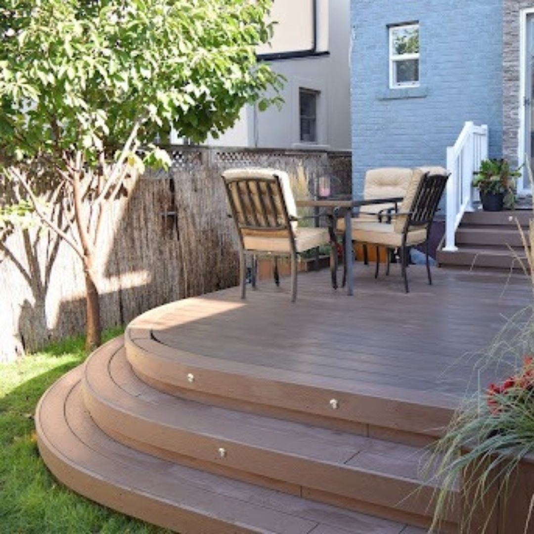 A curved brown composite deck with three stairs leads to a backyard patio area with a table and two chairs.