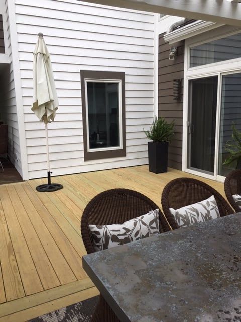 Patio deck with wicker chairs, closed umbrella, potted plant, and sliding glass doors beside a white house