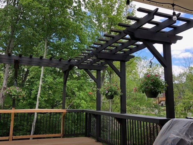 A dark wooden pergola built over a deck railing, with three hanging flower baskets and string lights against a tree line.