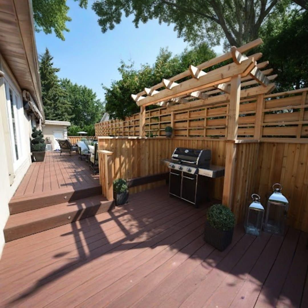 A wooden backyard deck with steps, a grill station, a privacy fence, and an overhead pergola on a sunny day.