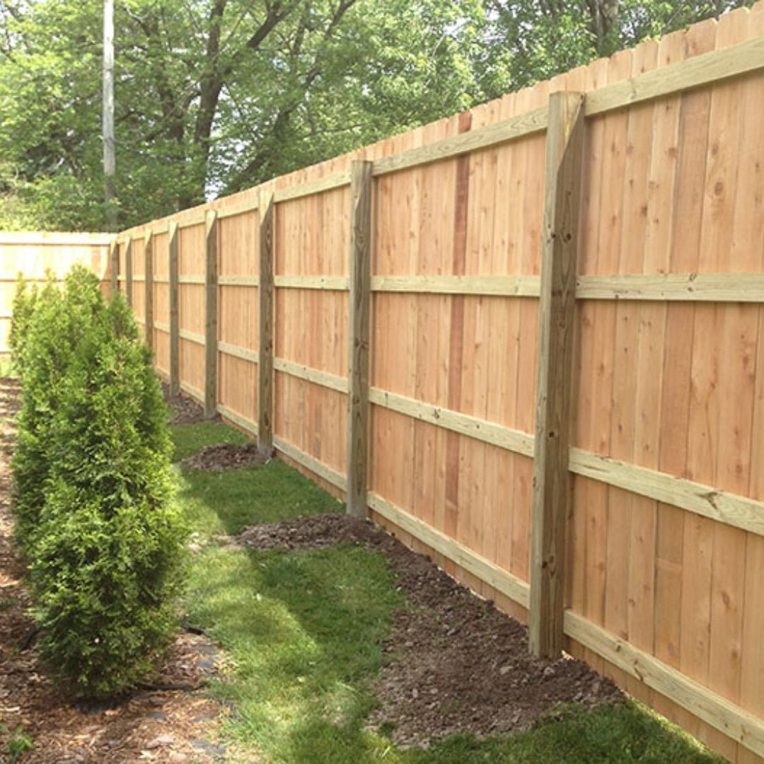 Wooden privacy fence along a narrow grassy path beside trees and shrubs.