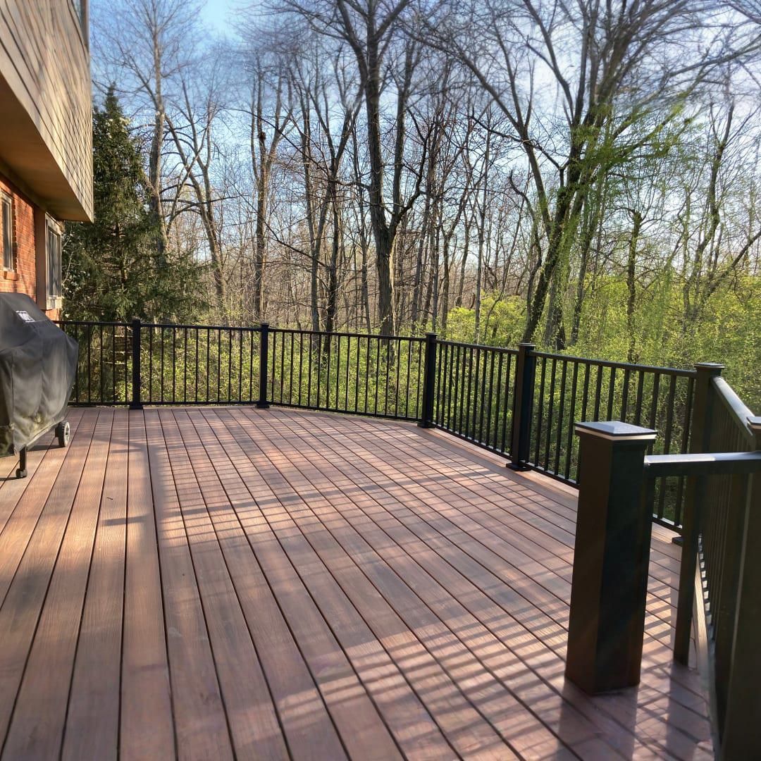 A wooden deck with black railings overlooks a dense, green treeline under a bright, sunny sky.