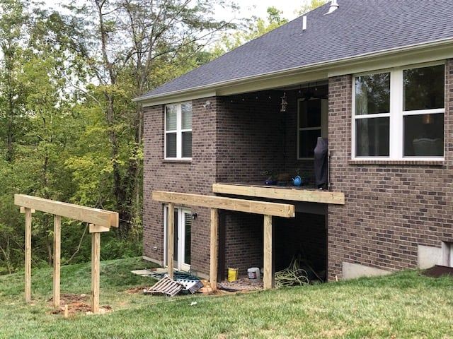 A multi-story brick house with a partially constructed wooden deck frame extending over a sloped, grassy backyard.