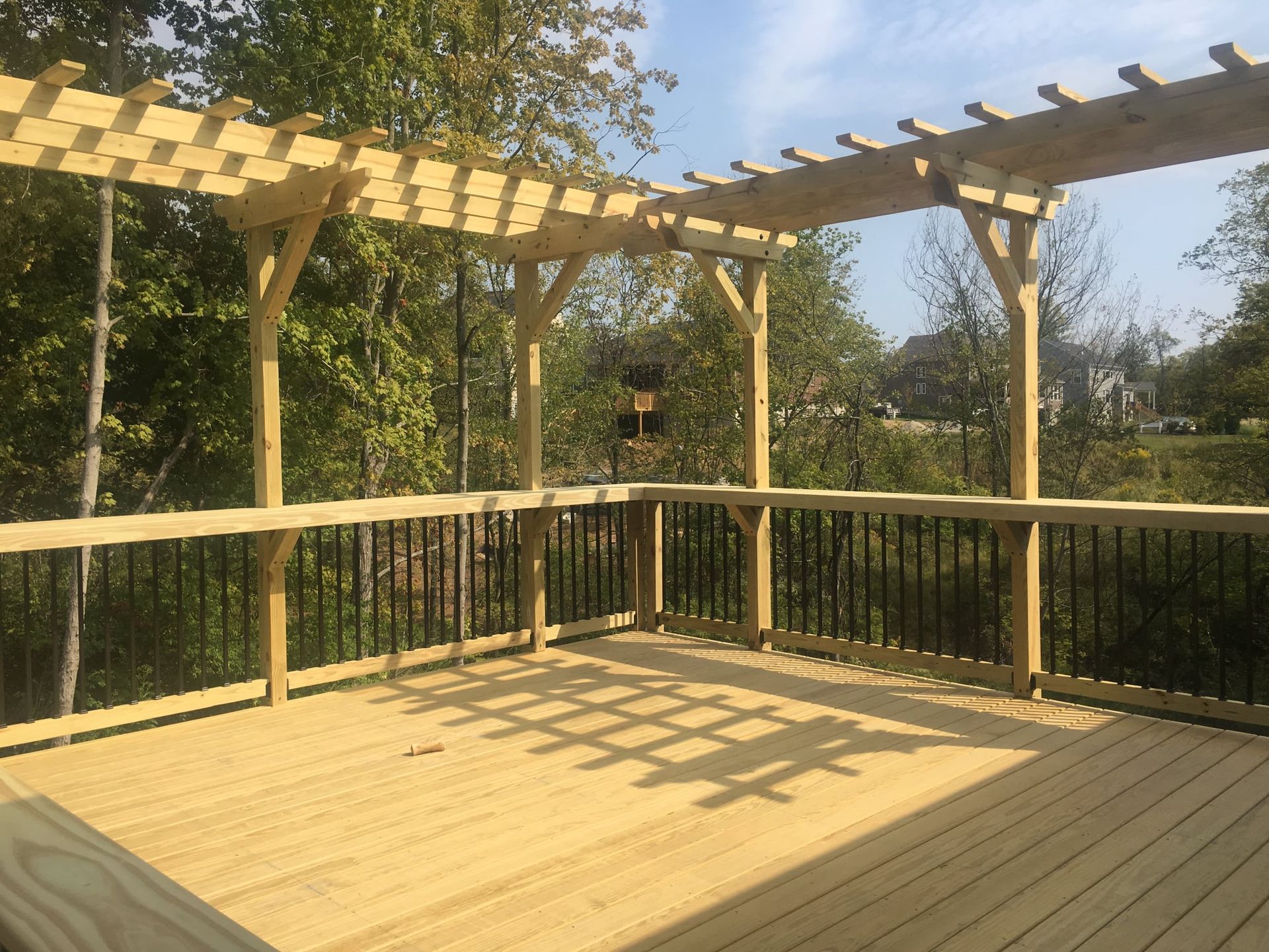 A wooden deck with a built-in railing bar and an overhead pergola, set against a backdrop of trees and a bright sky.