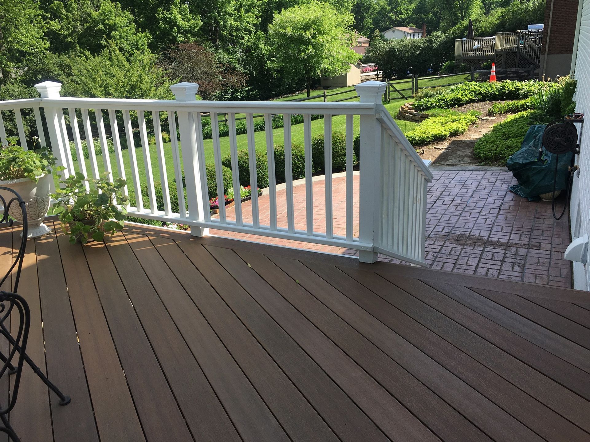 A dark wooden deck with white railing overlooks a backyard with a brick patio, manicured hedges, and lush green trees.