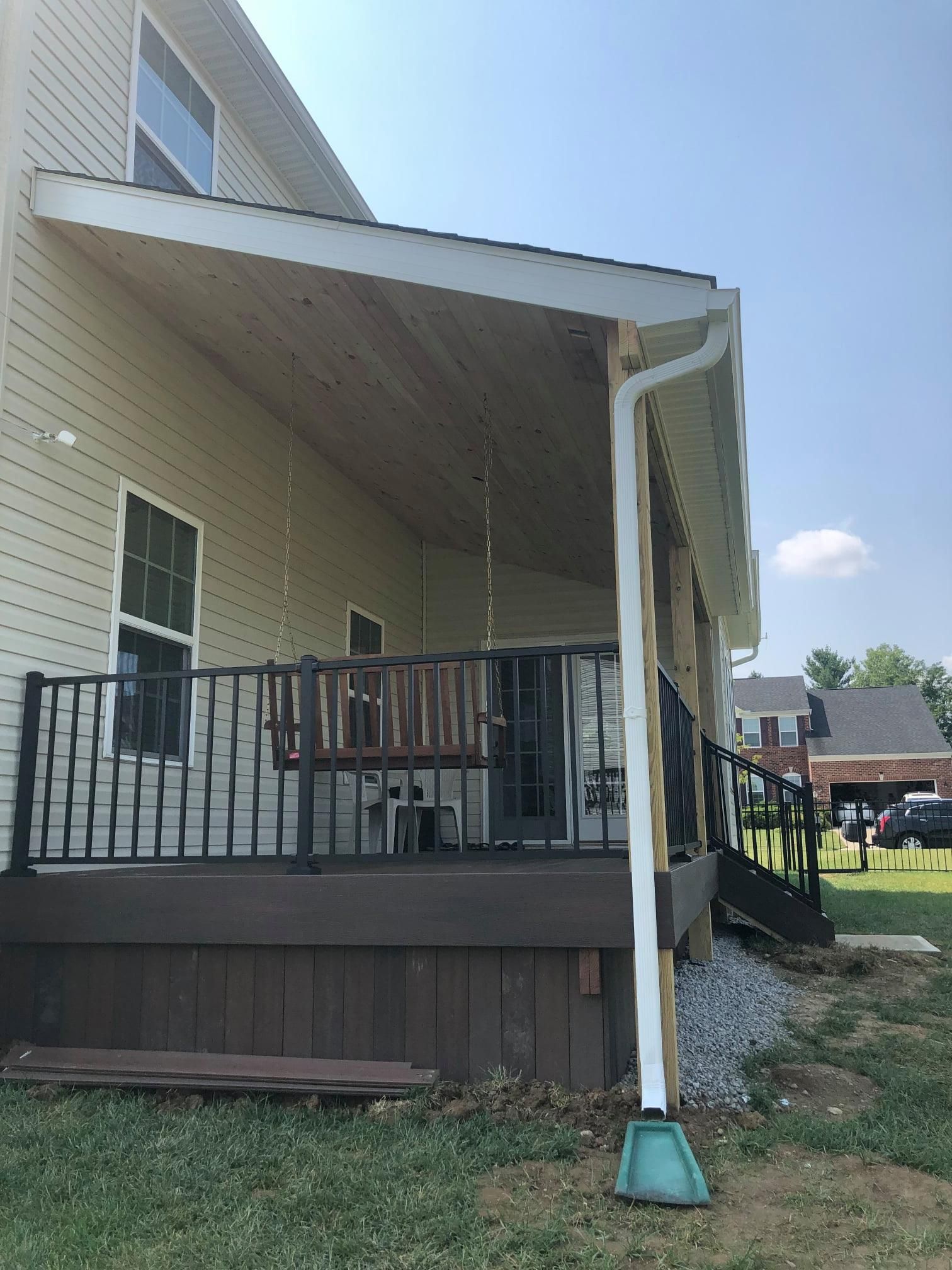A tan house with a covered wooden deck featuring a porch swing and black metal railings, alongside a white downspout.