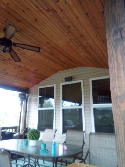A covered patio with a wooden ceiling, ceiling fan, outdoor dining set, and three windows on a light-colored siding wall.