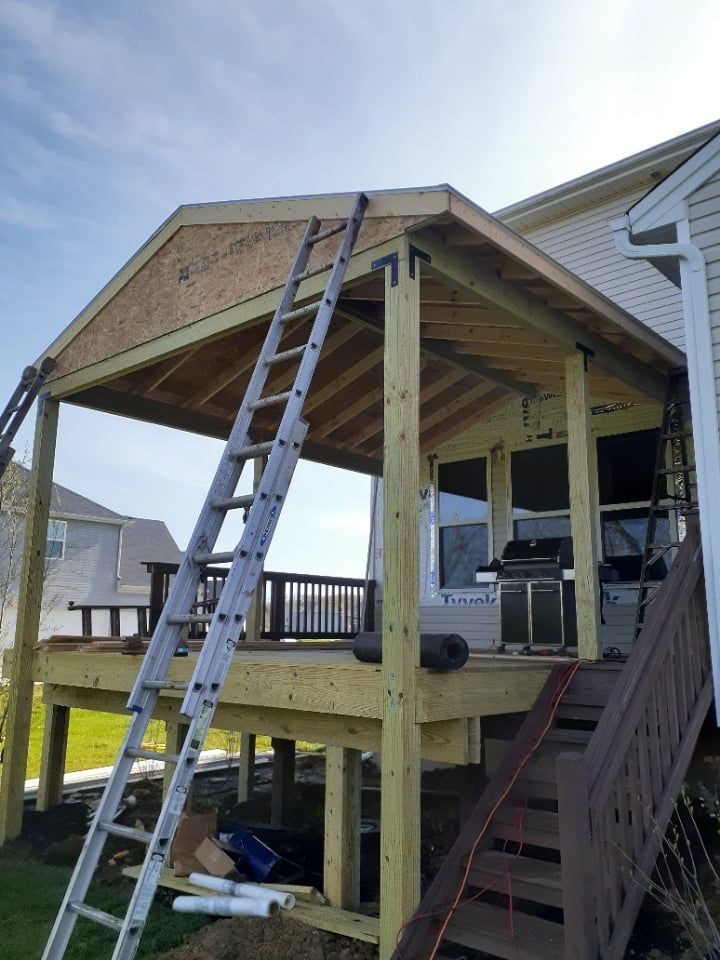 A wooden frame for a deck porch addition under construction, featuring an extended roof and a ladder leaning against it.