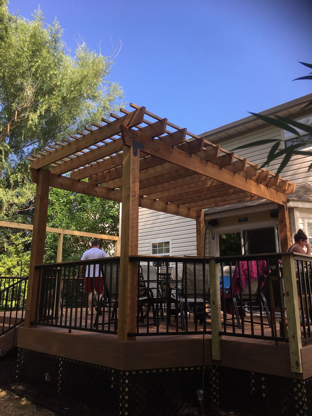 Wooden pergola-covered deck with railings in a backyard, beside a house and trees under a blue sky