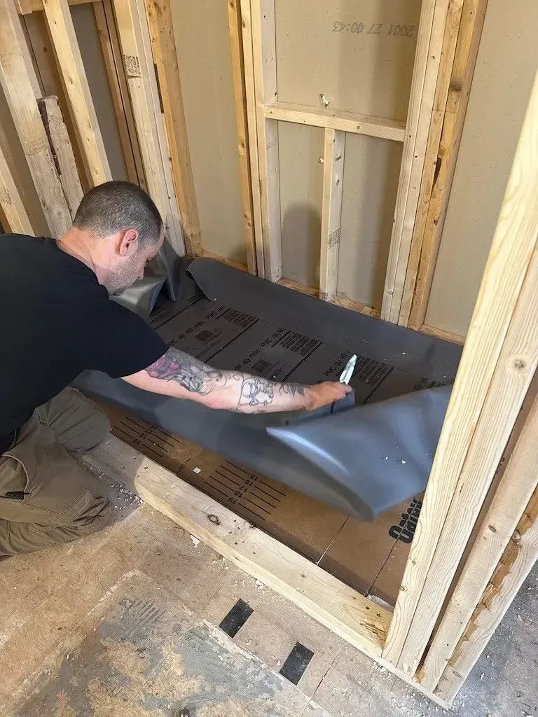 A person cuts a dark sheet inside a wooden shower frame. The person has tattoos and is wearing a black shirt.