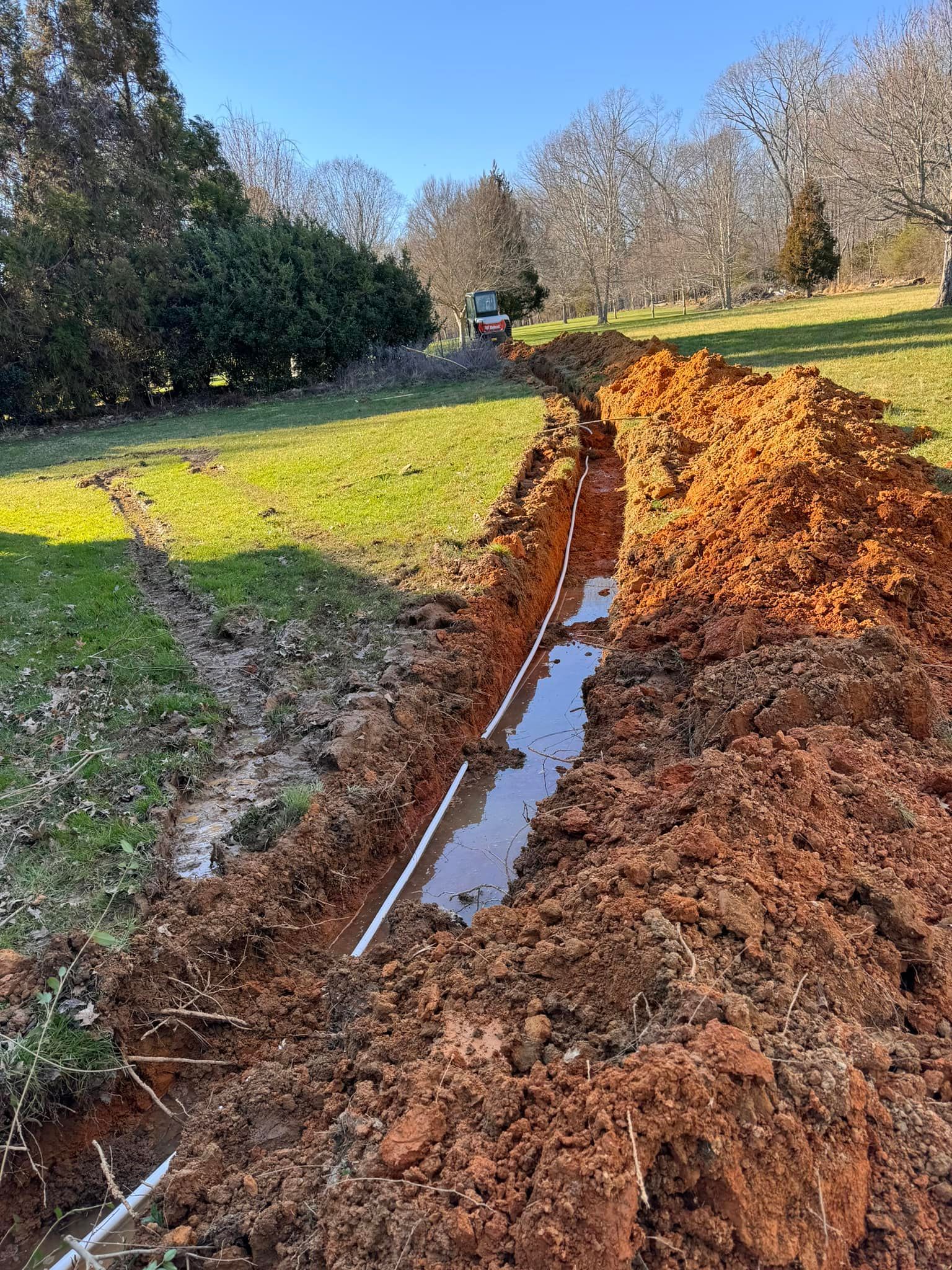 Dug trench in a grassy yard, containing a white pipe and water, with a small vehicle in the distance.
