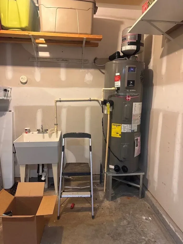 Garage interior with a water heater, utility sink, and a step ladder. A cardboard box is on the floor.