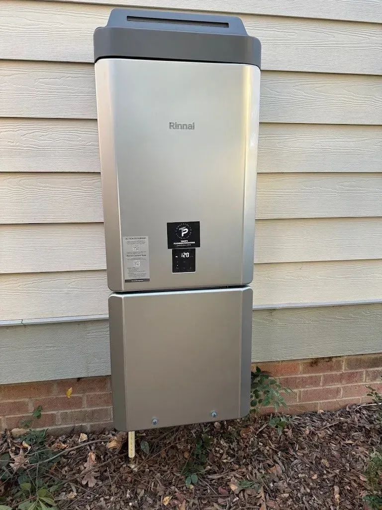 A silver Rinnai water heater mounted on a wooden building, with a brick foundation and some grass.
