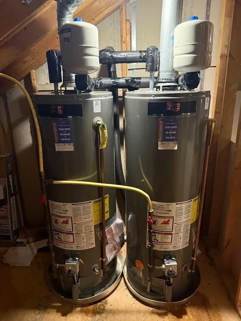 Two gray water heaters with white tanks in a wooden-walled attic.