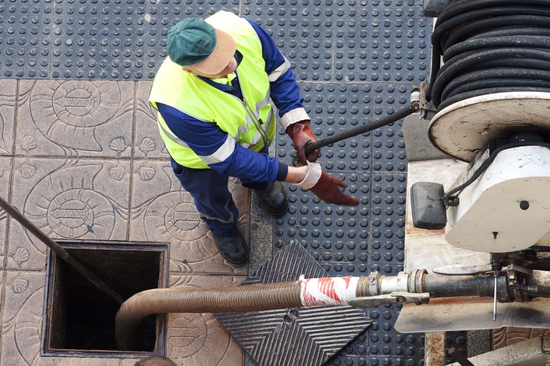 Man in safety vest working on a drain. He's inserting a hose into an open manhole, near a reel of hose.