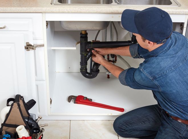 Plumber fixing pipes under a kitchen sink, wearing a blue cap and jeans, with a red wrench.