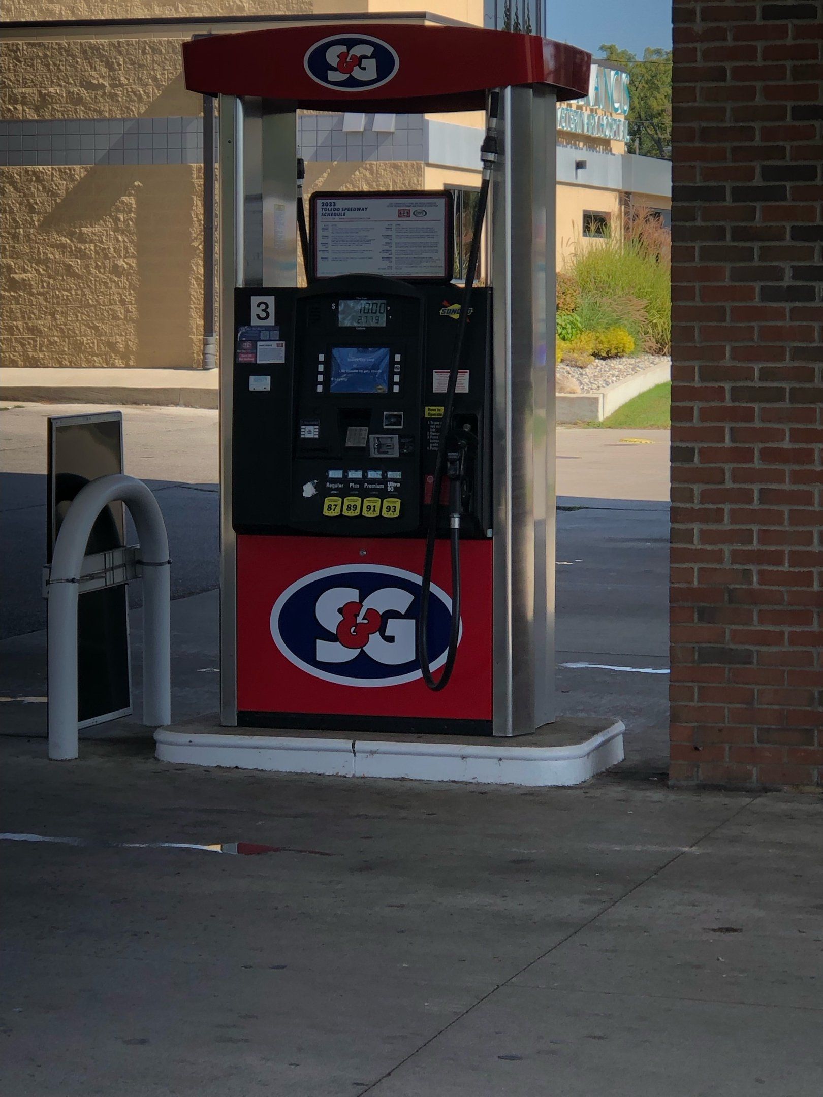 Gas pump with SG logo, standing on a concrete pad next to a brick building.