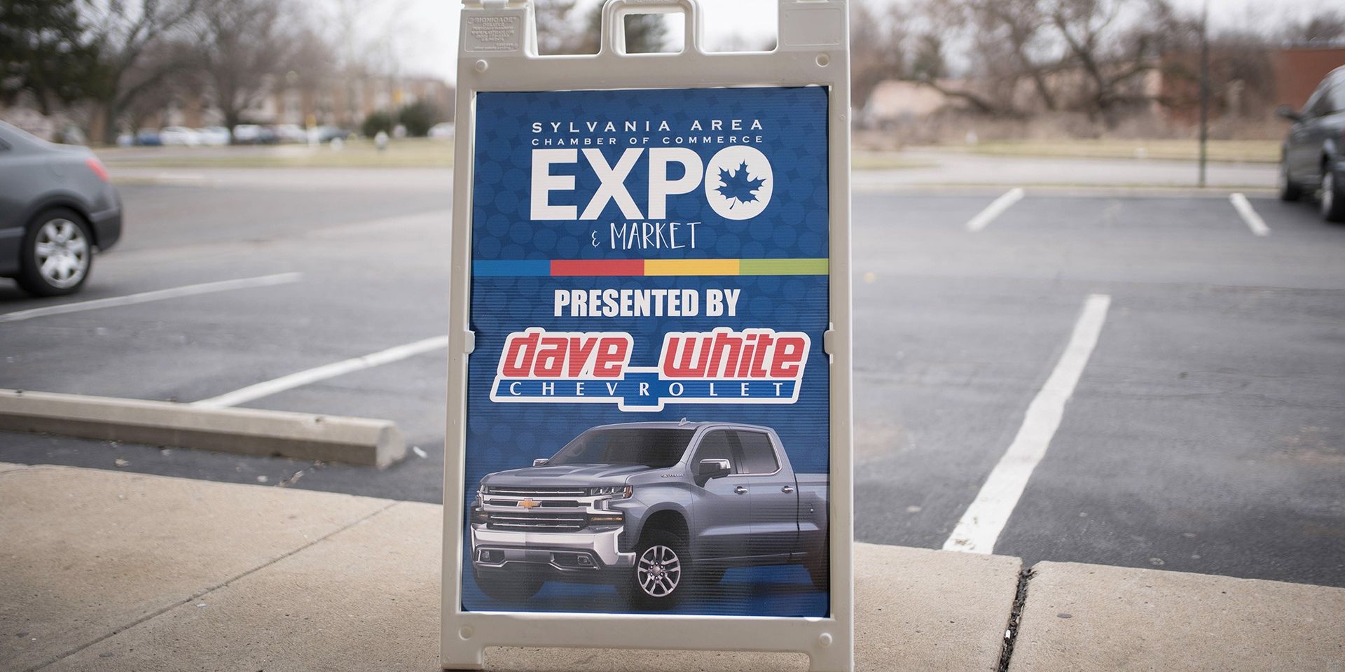 An A-frame sign in a parking lot promotes an expo presented by Dave White and featuring a truck.