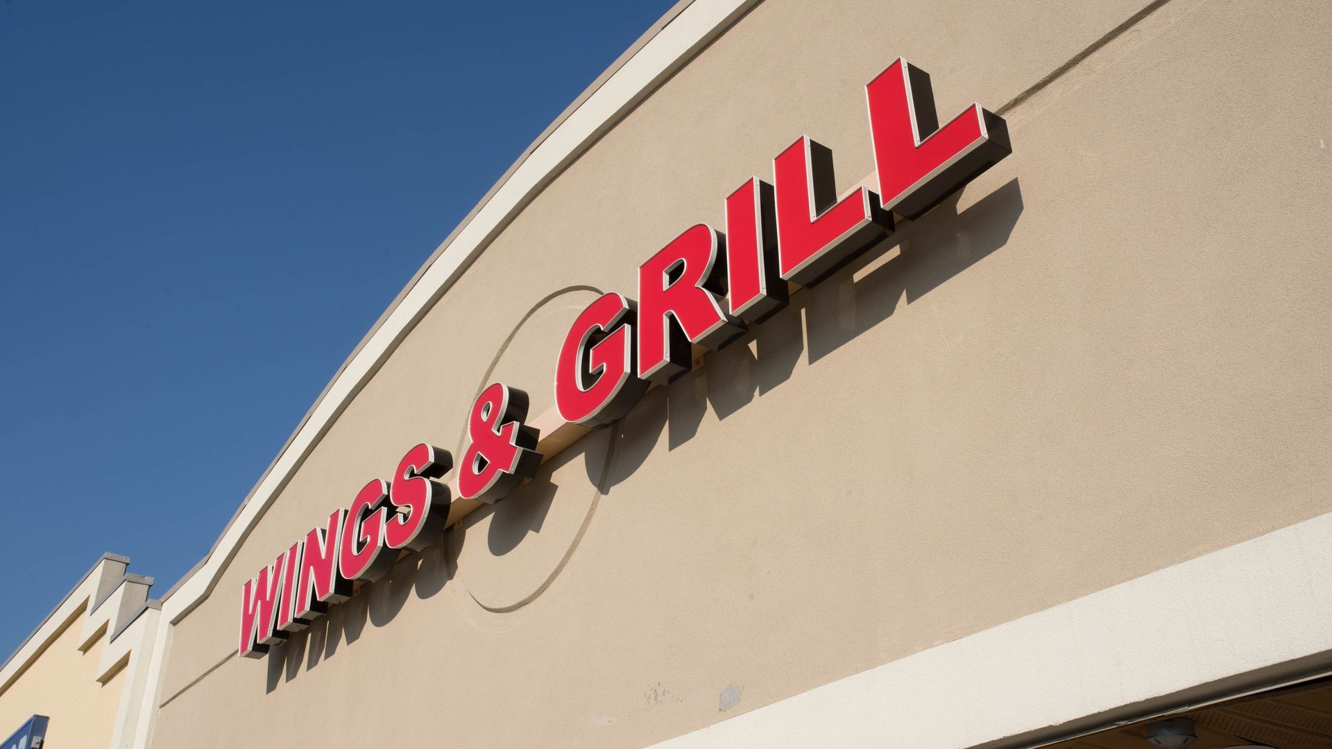 Wings & Grill restaurant sign, red letters on tan building under a blue sky.