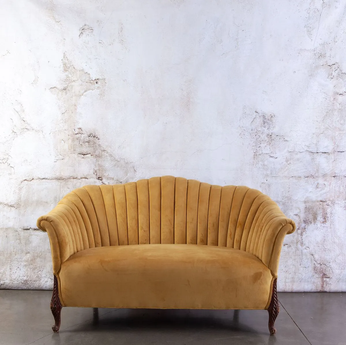Yellow velvet loveseat against a textured white wall, with dark wood legs.