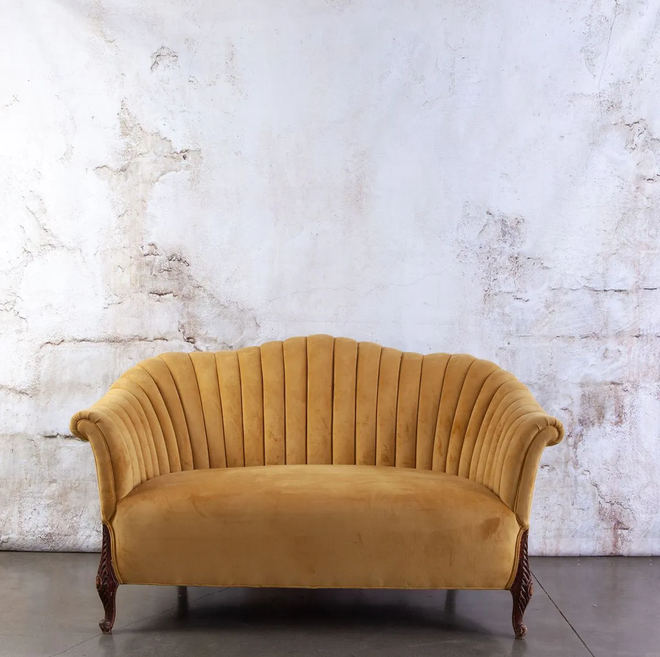 Yellow velvet loveseat against a textured white wall, with dark wood legs.