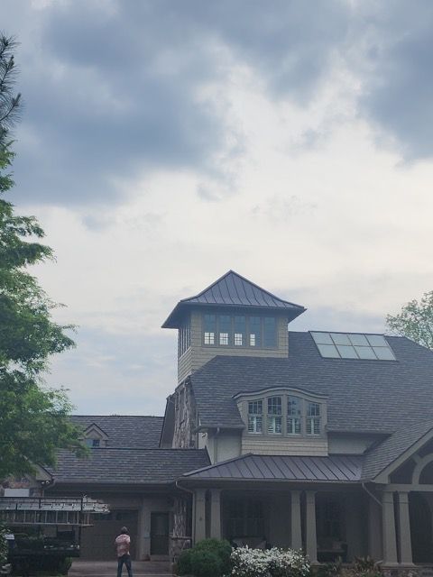 House with multiple rooflines and tower, cloudy sky. Man stands near garage door.