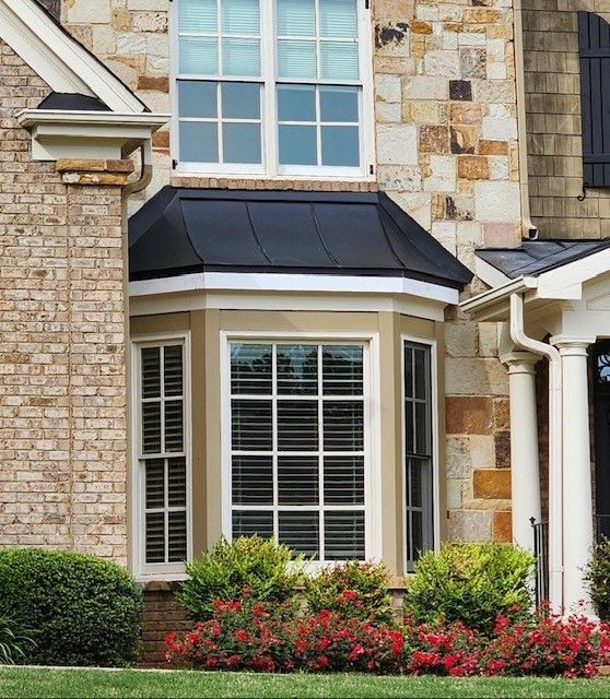 House exterior with stone and brick facade, bay window, black roof, and shrubs.
