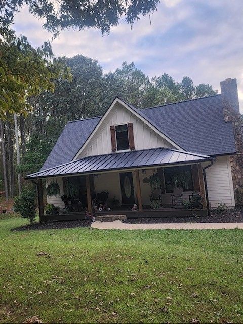 Cottage with a dark blue roof, porch, and chimney, surrounded by green grass and trees.