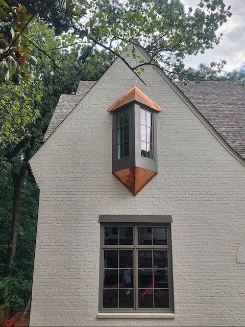 Brick building with copper bay window and brown-framed window, against a backdrop of trees.