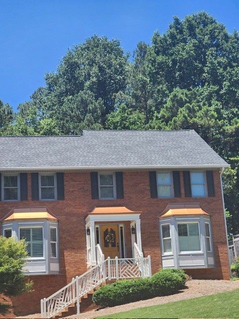 Two-story brick house with a gray roof and white trim, featuring bay windows and a yellow front door, with trees in the background.