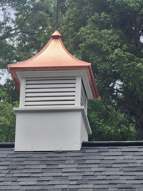Copper-topped roof vent on a dark shingle roof, white louvered sides, set against a backdrop of green trees.