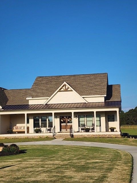 Beige farmhouse with a wraparound porch, brown roof, and blue sky.