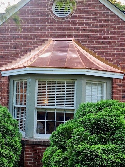 Copper roof over a bay window on a brick house. White trim and green shrubs.