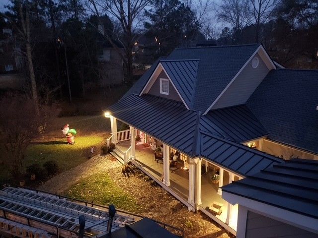 House with dark metal roof, lit porch and yard, trees in background, dusk.