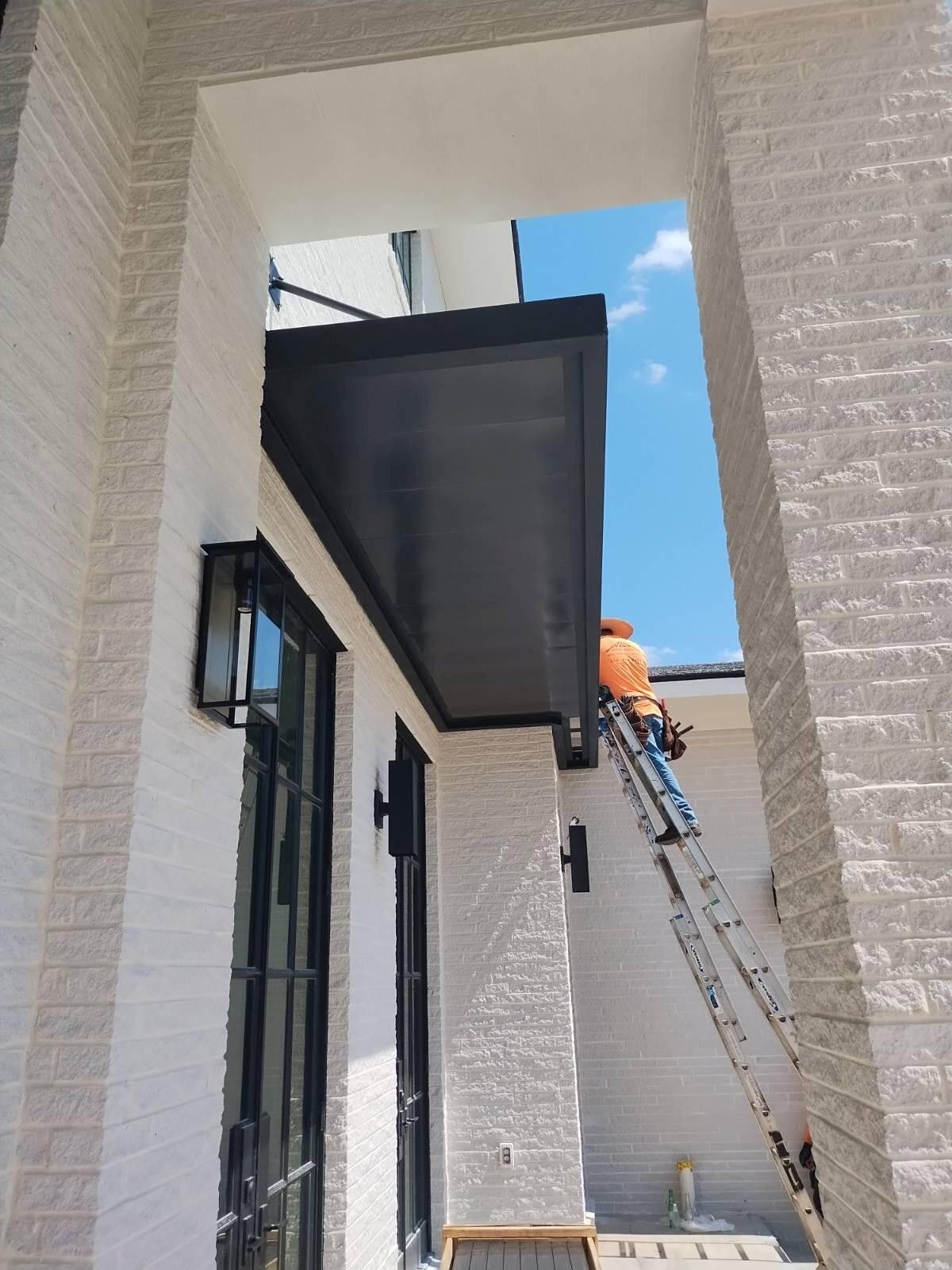 Worker on a ladder installing a black awning over a doorway, next to a brick wall under a blue sky.