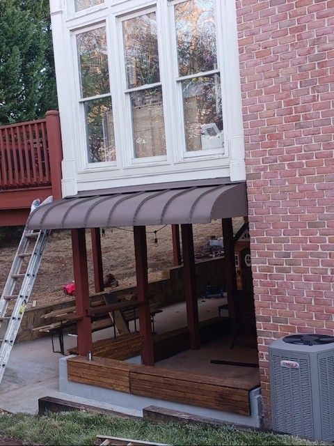 Brown awning over a deck under a building with windows, a brick wall and AC unit. A ladder leans against the deck.