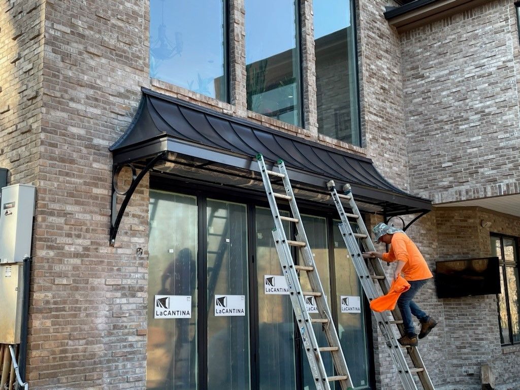 Man on ladder installing black metal awning over glass doors of a brick building.