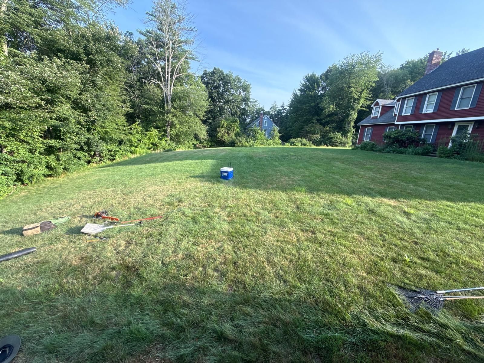 Backyard lawn beside a red house, with tools on the grass and trees along the edge.