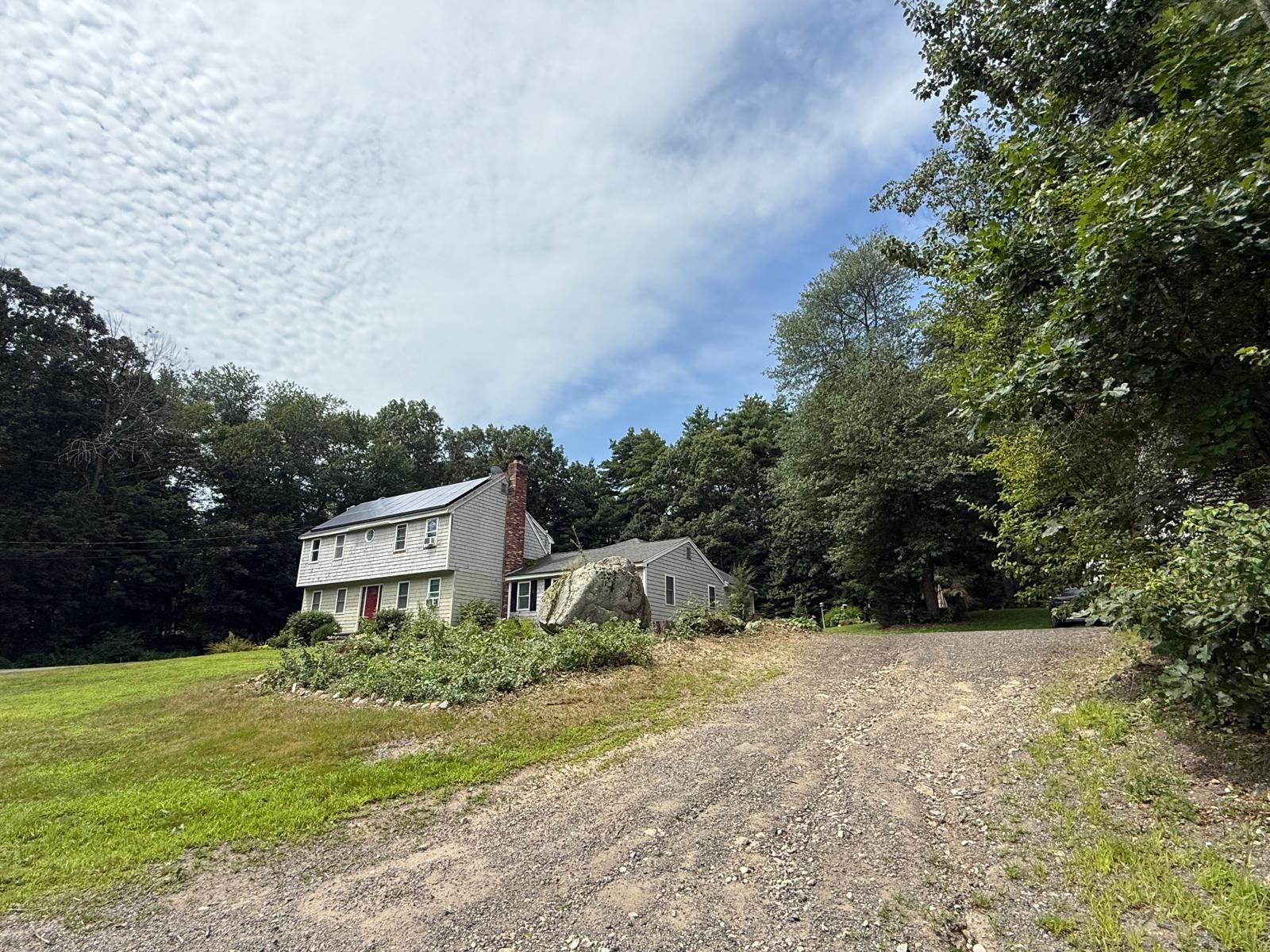 Rural stone house with gray roof beside a gravel driveway, surrounded by trees under a cloudy sky