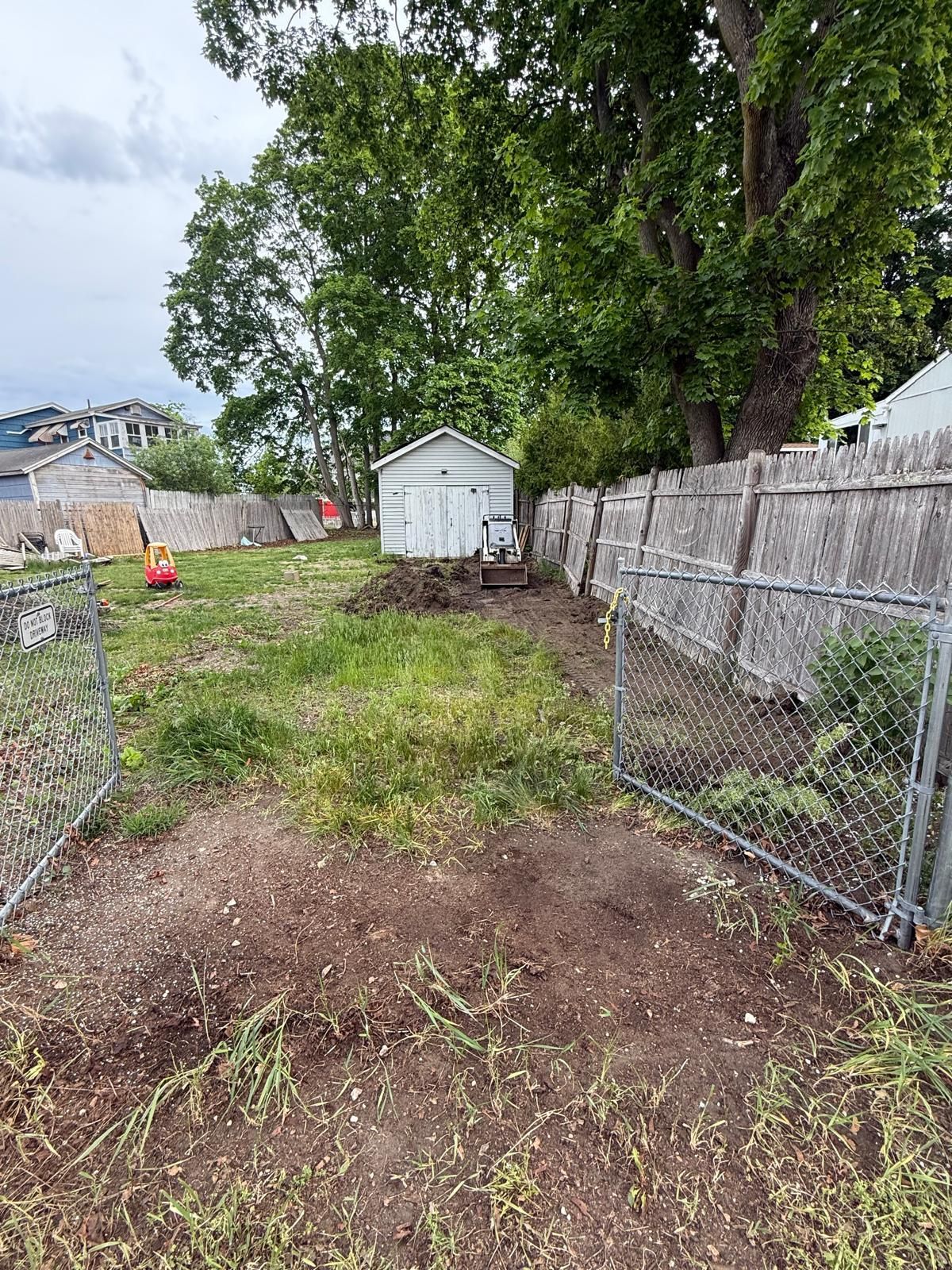 Chain-link fenced backyard with a small white shed, trees, and a dirt patch in the foreground.