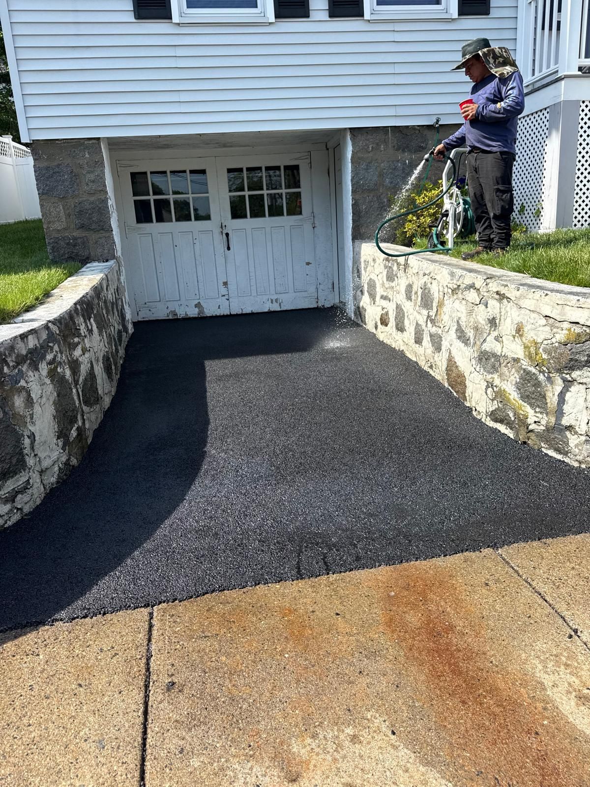 Freshly paved black driveway leading to a white garage beside stone retaining walls, with a worker standing on the right.
