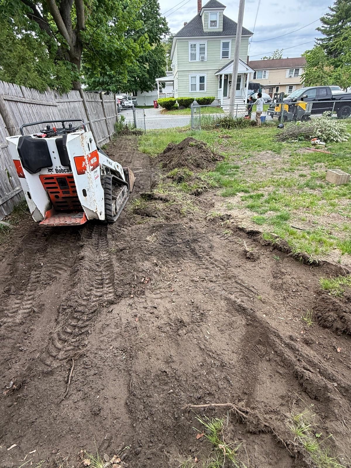 Small excavator with muddy tracks on a dirt lot near a house and fenced yard