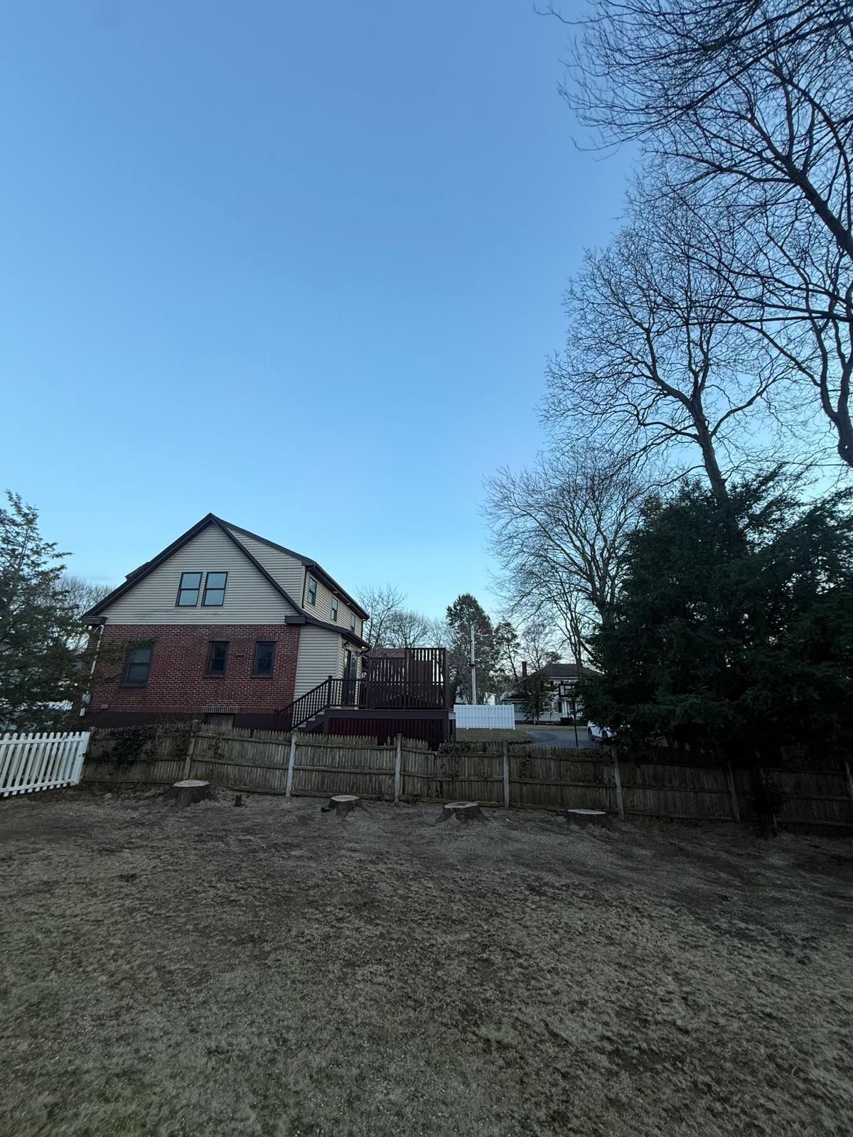Rural house with a red brick lower level, white upper gable, fenced yard, and leafless trees under a clear blue sky