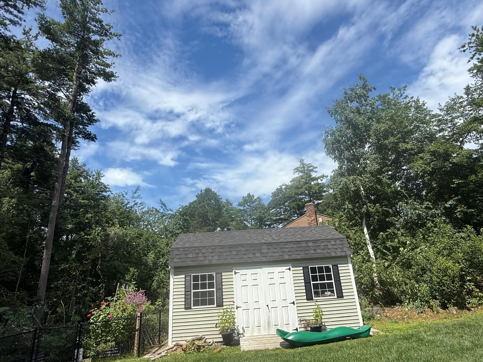 Small beige shed with white doors and green kayaks, set in a wooded yard under a cloudy blue sky.