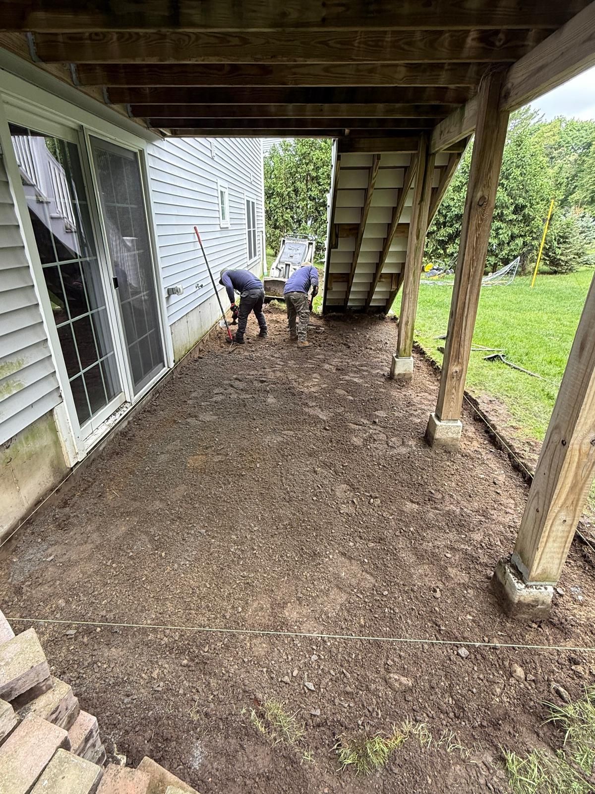 Two workers leveling dirt under a deck beside a white house and green lawn