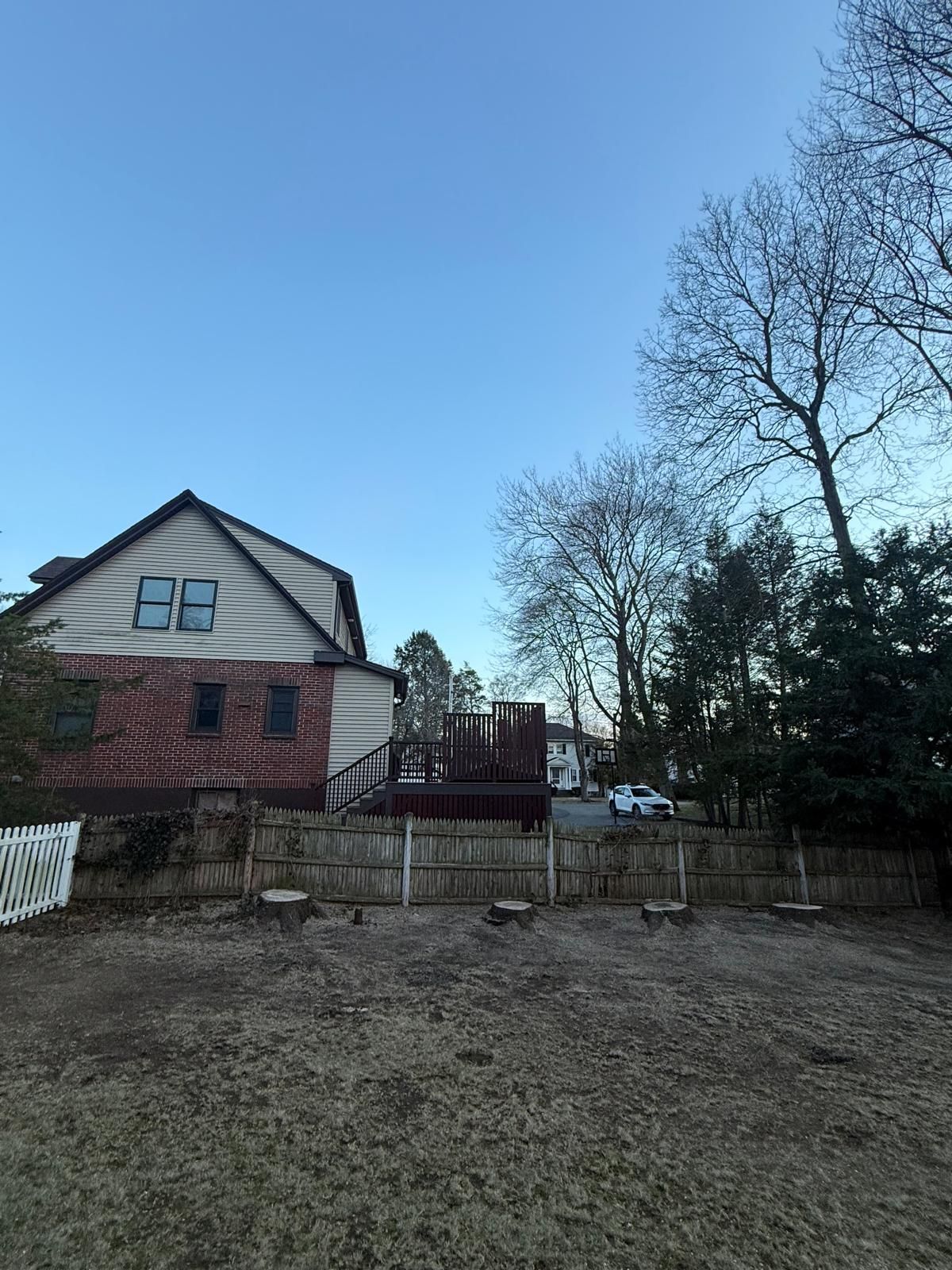 Backyard with a two-story house, wooden deck, fence, and leafless trees under a clear blue sky