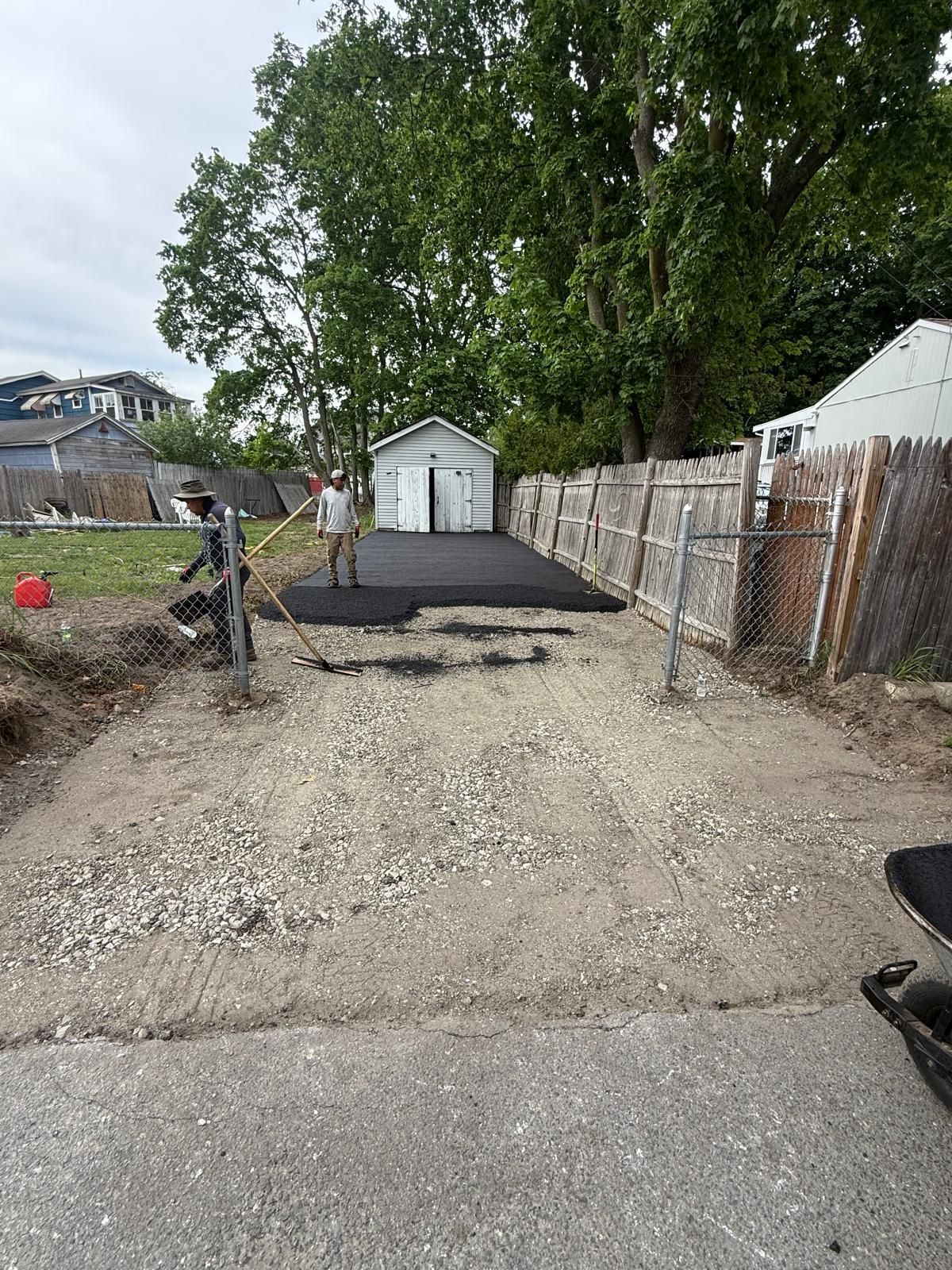 Gravel driveway under construction with workers, a black asphalt patch, trees, and a small white shed.
