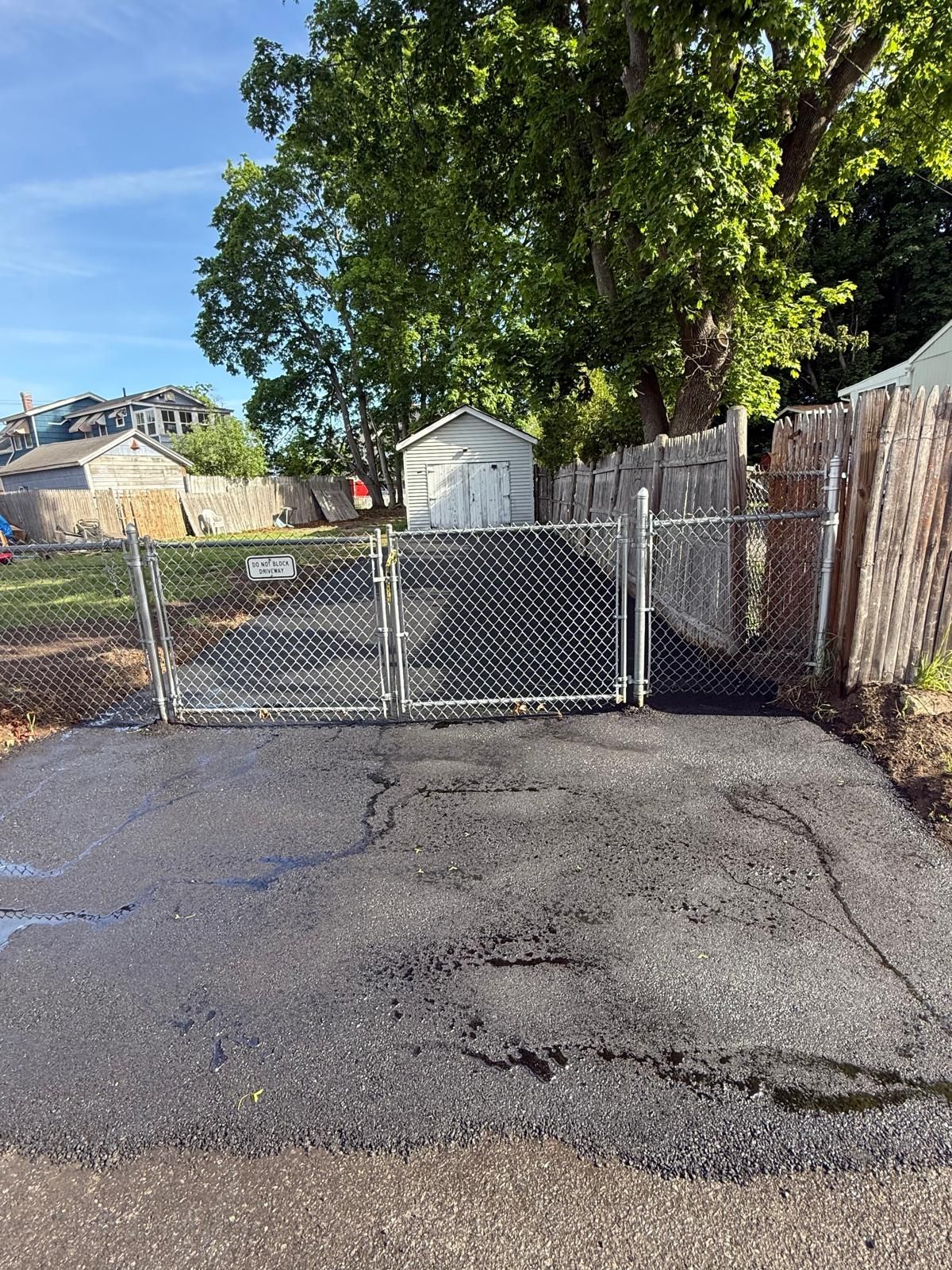 Chain-link gate blocking an empty cracked asphalt lot beside a tree and wooden fence under a blue sky