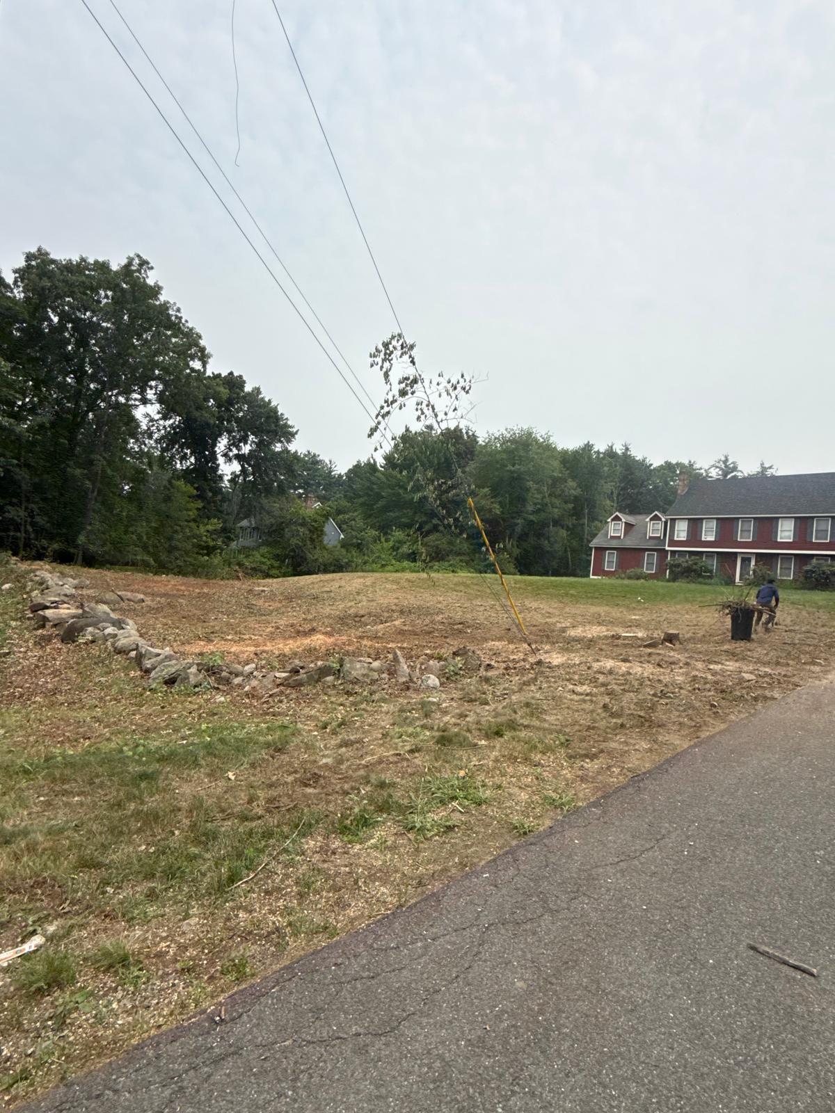 Rural roadside lot with cleared dirt, power lines overhead, trees, and a small house in the distance